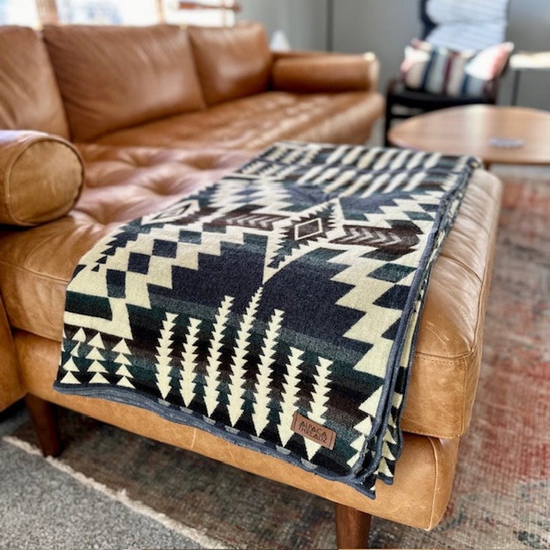 May include: A folded blanket with a geometric pattern in shades of blue, cream, and brown rests on a brown leather ottoman. The blanket has a dark blue border and a tag that reads "ALPACA THREADZ". The blanket is draped over a brown leather ottoman.