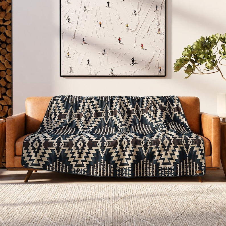 May include: A navy blue and beige geometric patterned throw blanket draped over a brown leather sofa. The blanket features a repeating zigzag design. A framed painting of skiers on a white background hangs above the sofa.