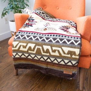 May include: A woven blanket with a geometric pattern in cream, brown, red, and gray tones is draped over an orange armchair. The blanket features a variety of shapes and lines, with a dark brown border and a small leather tag.