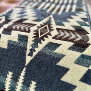 May include: Close-up of a textile with a geometric pattern in shades of blue, brown, and cream. The design features repeating shapes, including triangles, diamonds, and arrow-like motifs, creating a textured appearance. The fabric appears soft.