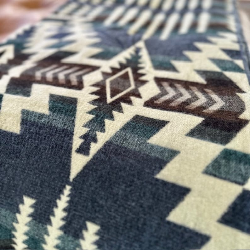 May include: Close-up of a textile with a geometric pattern in shades of blue, brown, and cream. The design features repeating shapes, including triangles, diamonds, and arrow-like motifs, creating a textured appearance. The fabric appears soft.