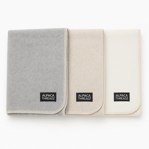 Könnte beinhalten: Drei gefaltete Decken in Grau-, Beige- und Weißtönen werden gezeigt. Jede Decke hat einen genähten Rand und ein rechteckiges Etikett mit der Aufschrift "ALPACA THREADZ". Die Decken sind ordentlich angeordnet und zeigen ihre weiche Textur.