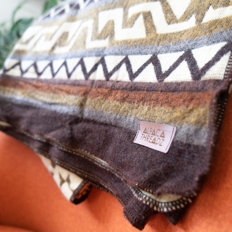 May include: A folded blanket with a geometric pattern in shades of brown, tan, and white. The blanket has a label that reads "ALPACA THREADZ". The blanket is made of a soft, textured material.