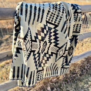 May include: A beige and black patterned blanket draped over a wooden fence. The blanket features geometric designs, including triangles, diamonds, and arrow-like shapes. The blanket's color palette includes beige, black, and dark green.