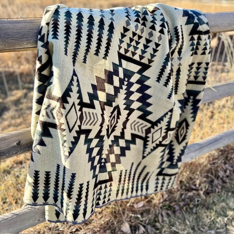 May include: A beige and black patterned blanket draped over a wooden fence. The blanket features geometric designs, including triangles, diamonds, and arrow-like shapes. The blanket's color palette includes beige, black, and dark green.