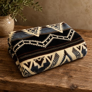 May include: Folded blanket with a Southwestern geometric pattern in black, cream, and blue. The blanket is made of a soft material and is folded neatly on a wooden surface. The design features triangles and zigzags.