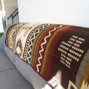 May include: A woven blanket with a geometric pattern in shades of brown, cream, red, and gold. The blanket is draped over a gray sofa, showcasing its intricate design and texture. The blanket's dimensions are not visible.