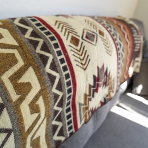 May include: A folded blanket with a geometric pattern in shades of cream, brown, red, and gray. The blanket is draped over a gray sofa, showcasing its intricate design and texture. The pattern includes zigzags, diamonds, and other shapes.