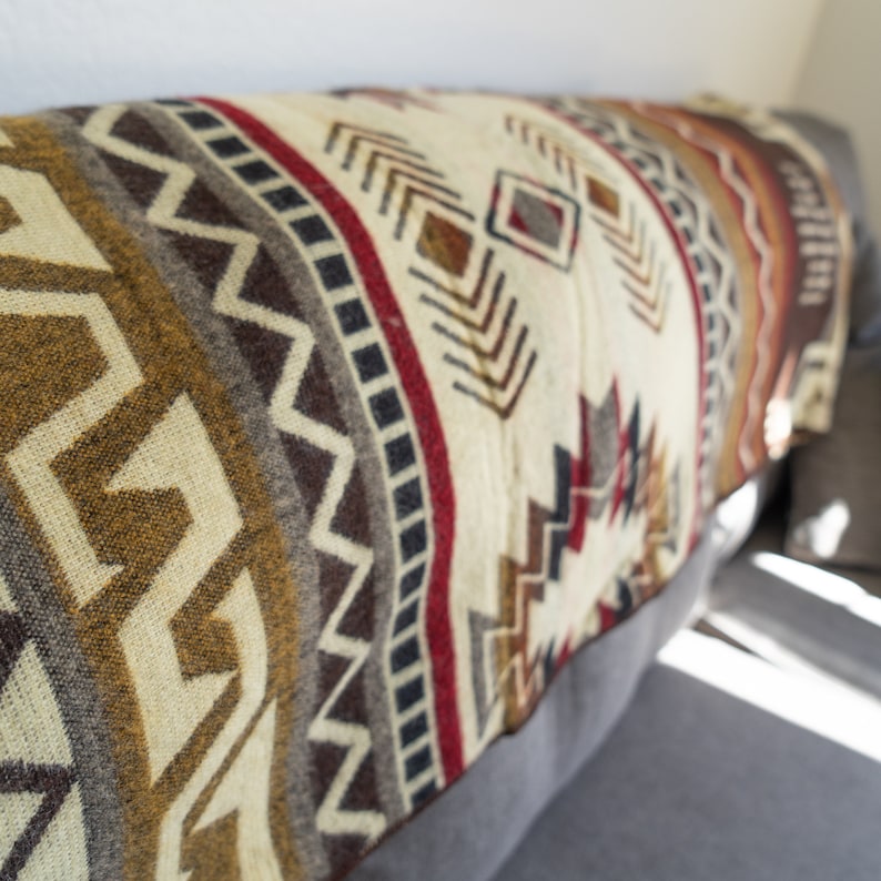 May include: A folded blanket with a geometric pattern in shades of cream, brown, red, and gray. The blanket is draped over a gray sofa, showcasing its intricate design and texture. The pattern includes zigzags, diamonds, and other shapes.