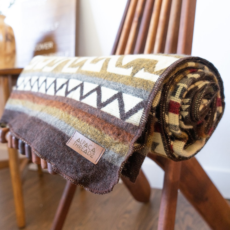 May include: A rolled-up blanket with a geometric pattern in shades of brown, cream, and tan, resting on a wooden chair. The blanket features a label that reads "ALPACA THREADZ". The blanket is made of a soft, woven material.