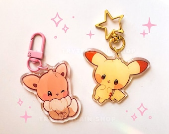 May include: Two adorable cartoon character keychains. The keychain on the left is a brown fox with a white tail. The keychain on the right is a yellow Pikachu with red cheeks. Both keychains are made of clear acrylic and have gold metal rings.