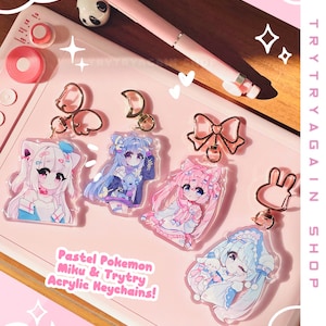May include: Pastel pink and blue anime character acrylic keychains with gold metal charms. The keychains feature characters with pastel pink, blue, and white hair and clothing. The text "Pastel Pokemon Miku & Trytry Acrylic Keychains!" is printed on the pink background.