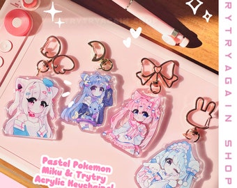 May include: Pastel pink and blue anime character acrylic keychains with gold metal charms. The keychains feature characters with pastel pink, blue, and white hair and clothing. The text "Pastel Pokemon Miku & Trytry Acrylic Keychains!" is printed on the pink background.