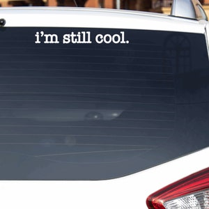 May include: A white car with a black rear window. The window has a white vinyl sticker that says "i'm still cool."