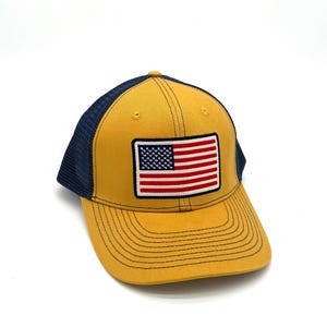 May include: Mustard yellow baseball cap with a navy blue mesh back and a curved brim. The cap features a rectangular patch with the American flag. The stitching is navy blue.