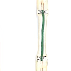 May include: A cream-colored, elongated fabric item with a central green stripe and red vertical stitching. Black stitching forms an X-shaped pattern at each end. The item appears to be a strap or a part of a larger textile.