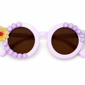 May include: Light purple round sunglasses featuring dark brown lenses. The left frame is decorated with white letter beads spelling BIG SISTER and a yellow flower. The right frame features white letter beads spelling ARIA and a light purple flower.