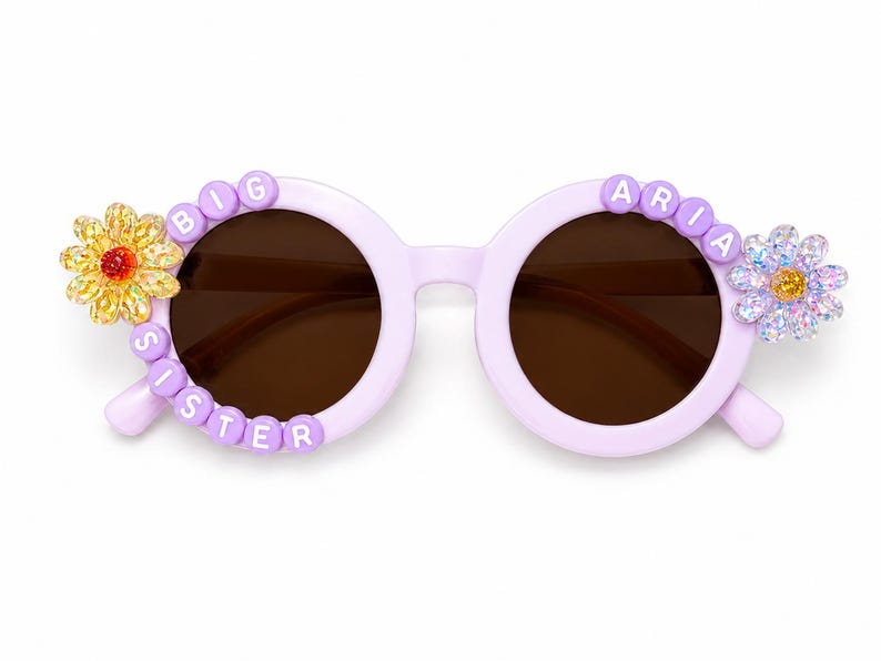 May include: Light purple round sunglasses featuring dark brown lenses. The left frame is decorated with white letter beads spelling BIG SISTER and a yellow flower. The right frame features white letter beads spelling ARIA and a light purple flower.