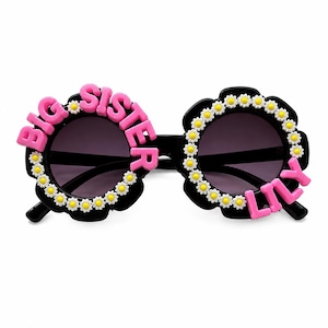 May include: Black sunglasses with round frames decorated with small white and yellow daisy flowers. The left lens frame features the pink text BIG SISTER, while the right lens frame features the pink text LILY, both attached to the outer edges.