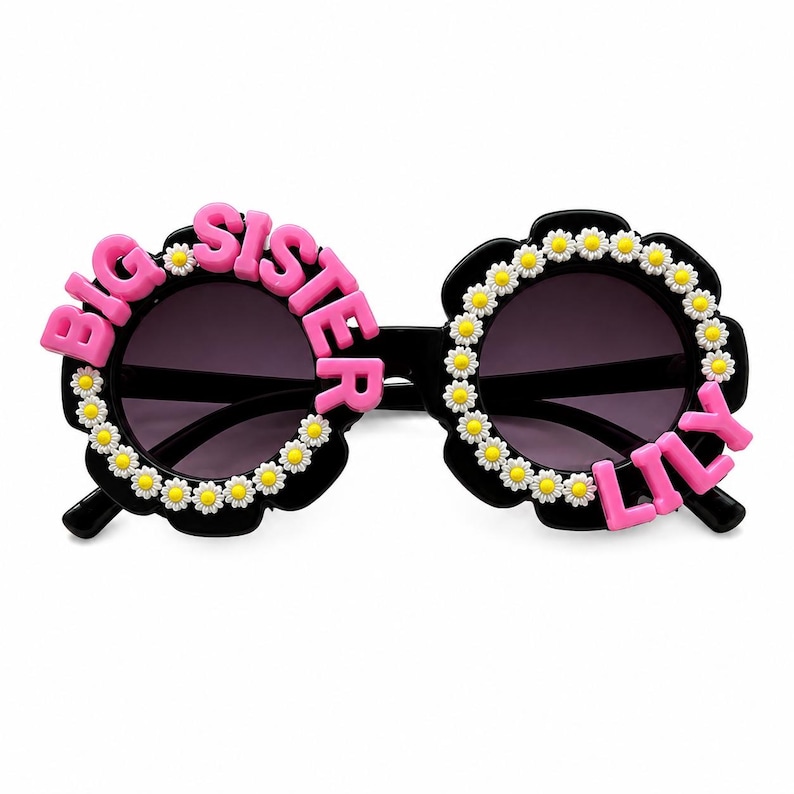 May include: Black sunglasses with round frames decorated with small white and yellow daisy flowers. The left lens frame features the pink text BIG SISTER, while the right lens frame features the pink text LILY, both attached to the outer edges.