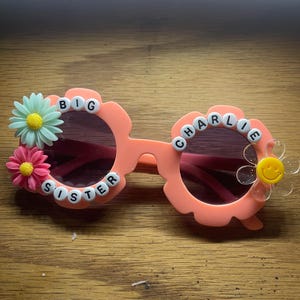 May include: Peach-colored flower-shaped sunglasses featuring white letter beads spelling BIG SISTER CHARLIE. The frames are decorated with a light green daisy, a pink daisy, and a clear flower with a yellow smiley face center on the right lens.