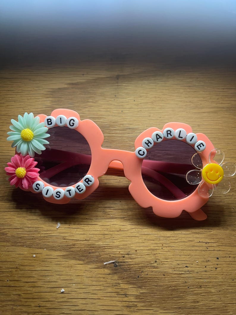 May include: Peach-colored flower-shaped sunglasses featuring white letter beads spelling BIG SISTER CHARLIE. The frames are decorated with a light green daisy, a pink daisy, and a clear flower with a yellow smiley face center on the right lens.