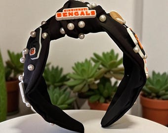 May include: Black knotted headband with Cincinnati Bengals logo and pearl accents. The headband features a football-themed design with orange and white details. The accessory is adorned with small, round pearls.