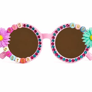 May include: Pink round-frame sunglasses decorated with alternating teal and magenta rhinestones. The left lens is bordered by beads spelling BIG SISTER, and the right lens features the letters ABI. A purple flower is on the left and a teal flower on the right.