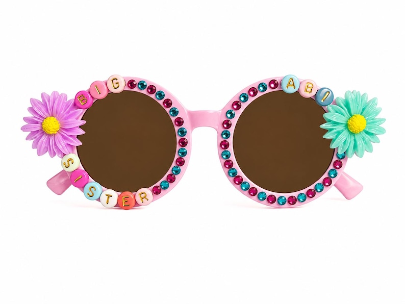May include: Pink round-frame sunglasses decorated with alternating teal and magenta rhinestones. The left lens is bordered by beads spelling BIG SISTER, and the right lens features the letters ABI. A purple flower is on the left and a teal flower on the right.