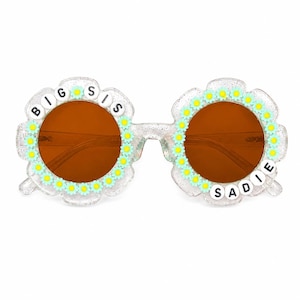 May include: Clear glittery flower-shaped sunglasses with brown lenses. The frames are decorated with small light blue and yellow daisy charms. The left lens is bordered by white beads spelling BIG SIS and the right lens features the name SADIE.