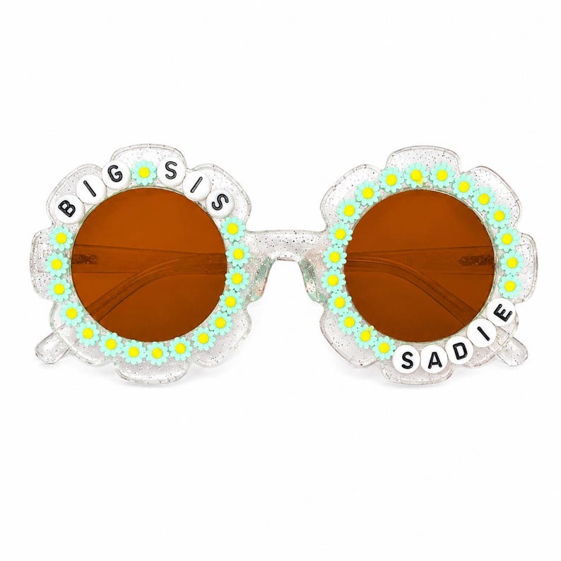 May include: Clear glittery flower-shaped sunglasses with brown lenses. The frames are decorated with small light blue and yellow daisy charms. The left lens is bordered by white beads spelling BIG SIS and the right lens features the name SADIE.
