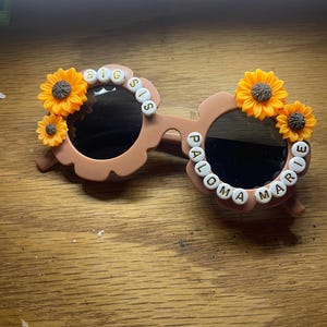 May include: Light brown flower-shaped sunglasses featuring yellow sunflower embellishments on the outer rims. The left lens frame is decorated with white letter beads spelling BIG SIS, while the right lens frame features the name PALOMA MARIE.