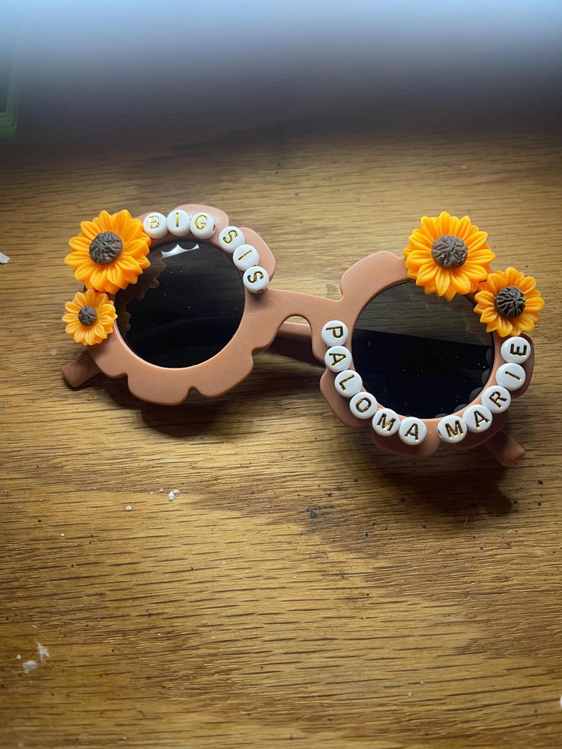 May include: Light brown flower-shaped sunglasses featuring yellow sunflower embellishments on the outer rims. The left lens frame is decorated with white letter beads spelling BIG SIS, while the right lens frame features the name PALOMA MARIE.