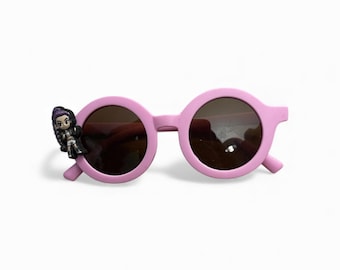 May include: Round sunglasses with a light pink plastic frame and dark tinted lenses. A small, three-dimensional figurine of a character with purple hair and a black outfit is attached to the outer edge of the left lens frame.