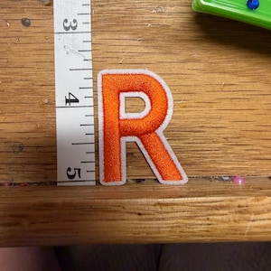 May include: An orange embroidered letter R patch with a white border. The patch is positioned next to a white measuring tape, showing the letter has a height of approximately 2 inches from the top to the bottom of the embroidery.
