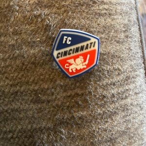 May include: A blue and red shield-shaped pin with the text "FC CINCINNATI" and a white lion with a red tail and a blue sword.