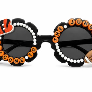 Op de afbeelding: Zwarte zonnebril met een rond montuur en een geschulpte rand, versierd met witte kralen en oranje letterkralen die "WELCOME TO THE JUNGLE" spellen. Het linker montuur heeft een gestreept logo en het rechter montuur bevat een football.