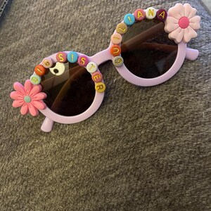 May include: Light purple round sunglasses featuring colorful letter beads spelling BIG SISTER on the left lens and GIULIANA on the right lens, accented with pink flower embellishments on the outer corners of the frames and dark tinted lenses.