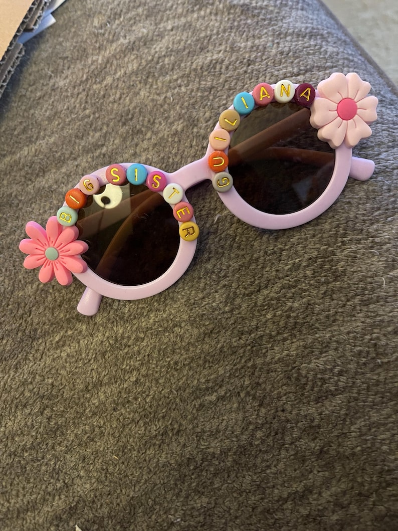 May include: Light purple round sunglasses featuring colorful letter beads spelling BIG SISTER on the left lens and GIULIANA on the right lens, accented with pink flower embellishments on the outer corners of the frames and dark tinted lenses.