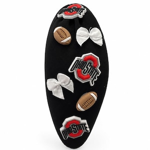Könnte beinhalten: Ein schwarzes Stirnband, verziert mit drei Ohio State University-Logos, zwei braunen Anhängern in Football-Form und zwei weißen Stoffschleifen. Die Anhänger und Logos sind auf der Oberfläche des mit schwarzem Stoff bezogenen Stirnbandes angeordnet.