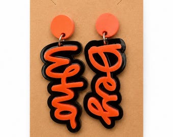 May include: A pair of dangle earrings featuring orange circular studs and black acrylic pendants with the orange text "Hey" and "Why" in a cursive script. The earrings are attached to a rectangular brown card with a circular hole at the top.