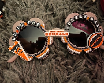 May include: Unique sunglasses with a floral frame design. The sunglasses feature the text "UR IN THE JUNGLE BABY" and "CINCINNATI BENGALS" with a football graphic. The frames are decorated with black, white, and orange embellishments.