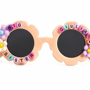 May include: Peach-colored flower-shaped sunglasses with dark lenses. The frames are decorated with colorful plastic beads spelling BIG SISTER on the left and GIULIANA on the right, accented with small purple, white, and yellow flower charms.