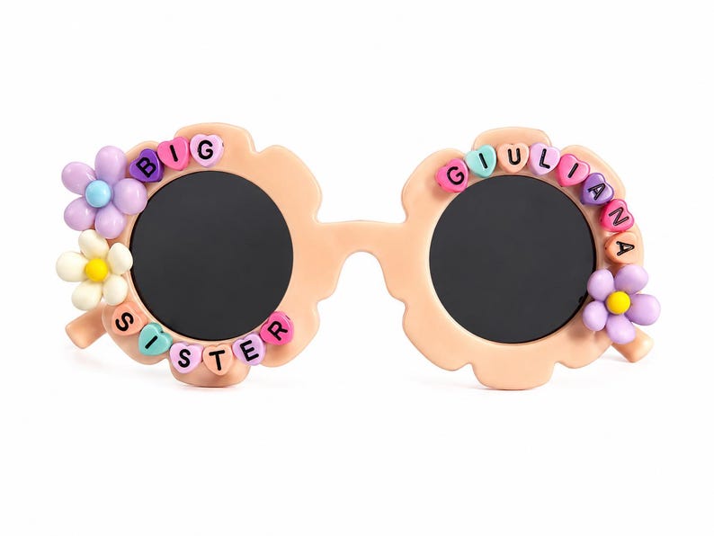 May include: Peach-colored flower-shaped sunglasses with dark lenses. The frames are decorated with colorful plastic beads spelling BIG SISTER on the left and GIULIANA on the right, accented with small purple, white, and yellow flower charms.