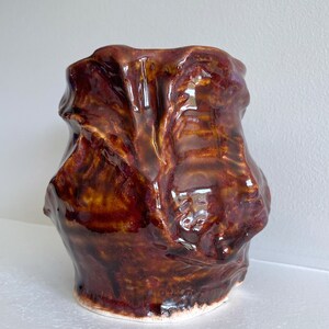 Puede incluir: Un jarrón de cerámica hecho a mano y brillante con una forma orgánica única. El jarrón presenta un rico esmalte marrón oscuro con toques de oro y rojo, creando una apariencia texturizada y escultural. La base es blanca.