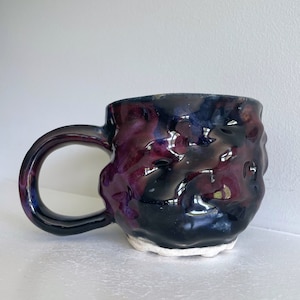 Puede incluir: Una taza de cerámica hecha a mano con una superficie texturizada. La taza presenta una combinación de colores oscuros con tonos de negro, morado y azul. El asa es oscura y la base tiene un aspecto blanco sin esmaltar. La taza está diseñada para bebidas calientes.