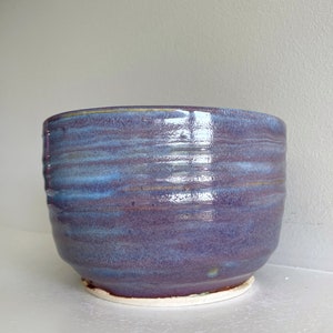 May include: A handmade ceramic bowl with a glossy, layered glaze in shades of purple and blue. The bowl has a rounded shape with a slightly flared rim and a white base. The bowl is suitable for serving food or as a decorative piece.