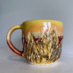 May include: A ceramic mug with a textured mountain design. The mug has a yellow, orange, and brown colour scheme. The handle is brown. The mug is on a white surface.