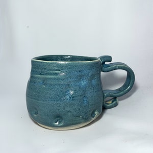 Peut inclure: Mug en céramique fait à la main, de couleur bleu-vert moucheté. Il présente une anse originale et une surface texturée avec de petites indentations. L'intérieur est bleu foncé. Idéal pour les boissons chaudes.