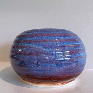 May include: A handcrafted ceramic bowl with a rounded shape. The bowl features a glossy finish with horizontal bands of blue and red-brown hues. The base is a lighter brown color. This decorative bowl is suitable for home decor.